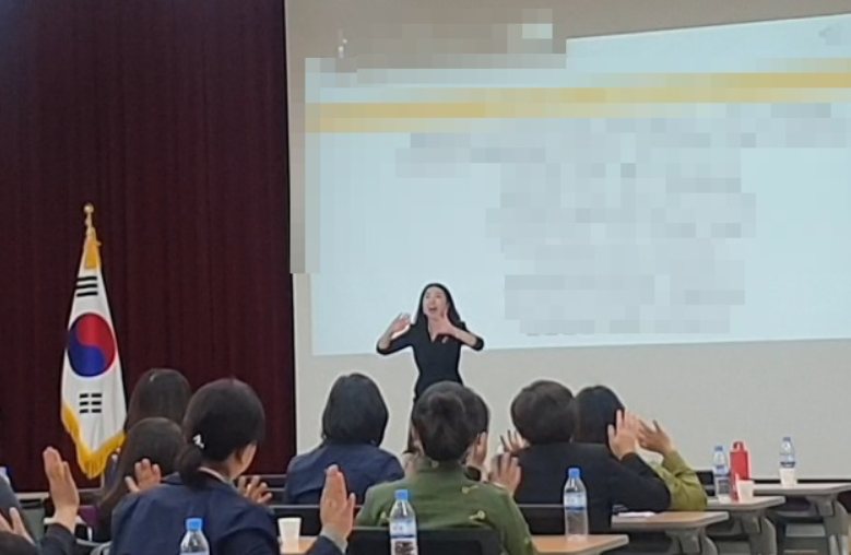 태극기가 놓인 기관 강당에서 직원들과 함께 박수 치며 강의하는 고아라 강사